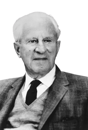 Black and white portrait of an elderly man with short, white hair, dressed in a suit jacket over a sweater and tie. He is seated in front of a bookshelf, gazing slightly to the side with a thoughtful expression. His formal attire and the background suggest an intellectual environment, conveying a sense of wisdom and scholarly presence.