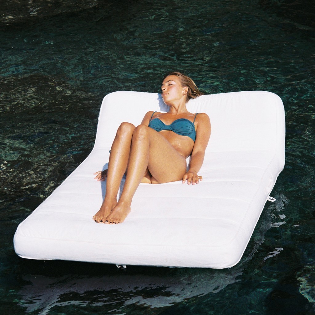 Woman sunbathing on a luxury all-white pool float floating in deep turquoise water