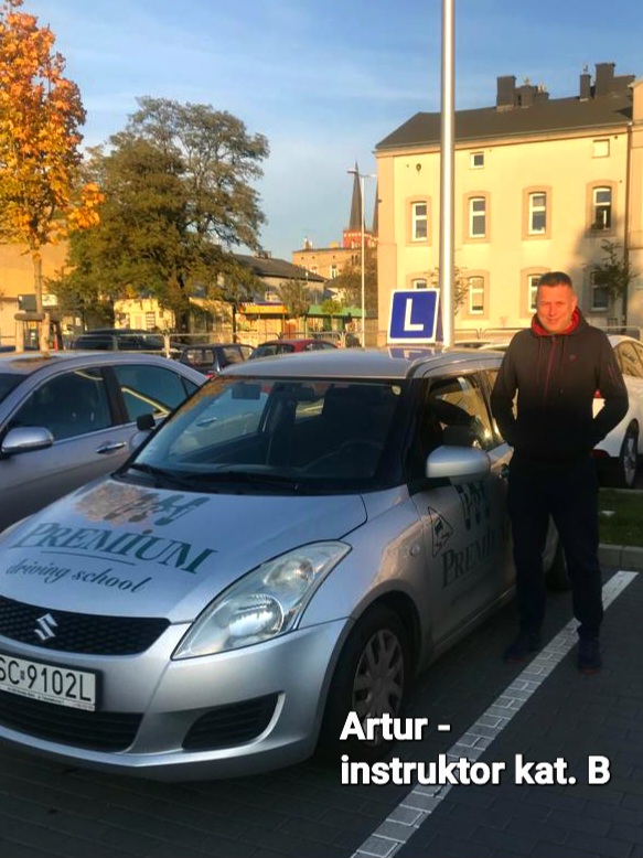 Artur – Instruktor pozujący obok srebrnego Suzuki Swift z logiem szkoły jazdy.