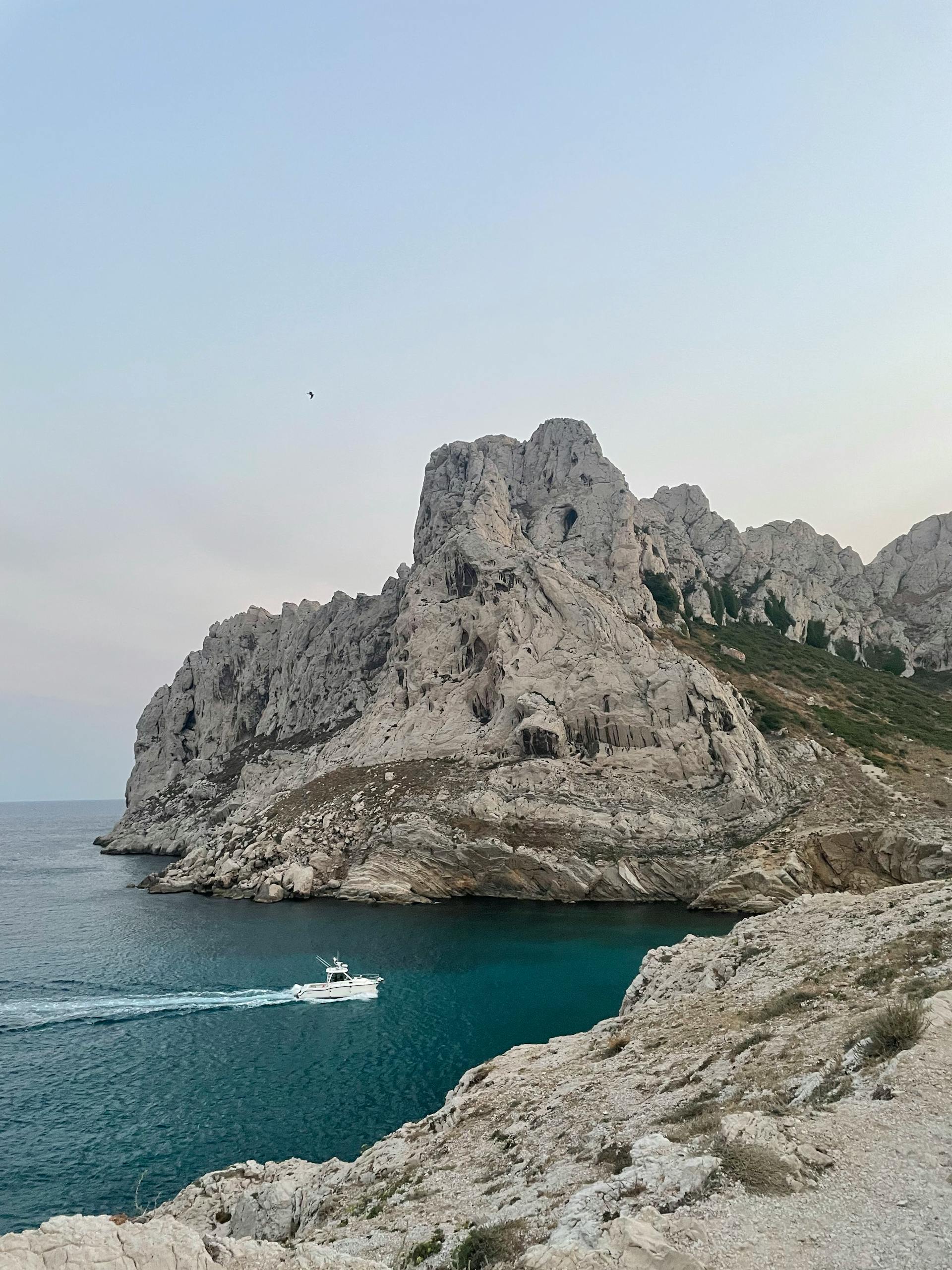 A small white motorboat cruises on turquoise water, leaving a wake, next to a massive, craggy white rock cliff formation under a hazy sky.