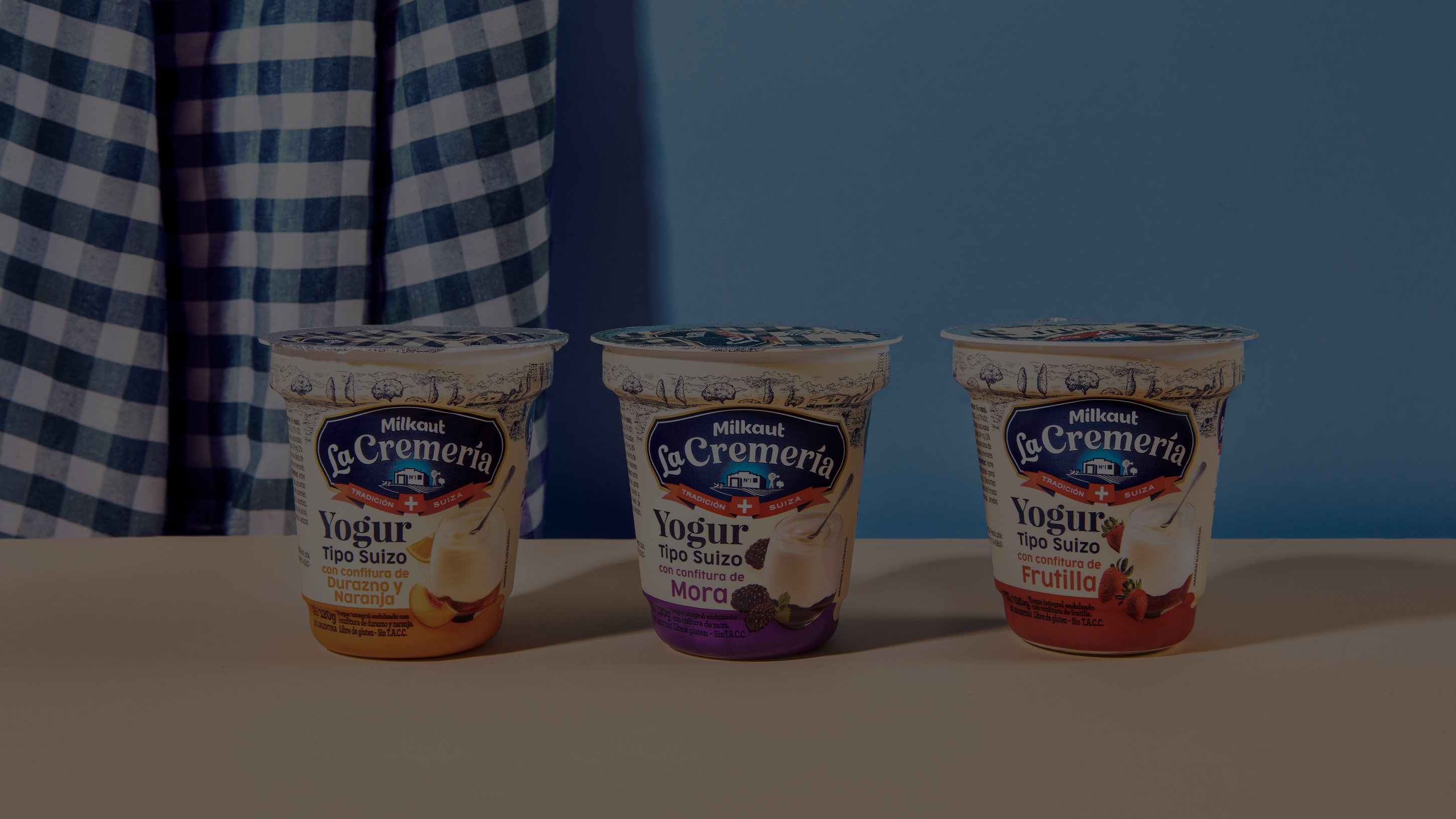 Three yogurt containers of different flavors—banana, blackberry, and strawberry—are arranged on a beige surface. A blue gingham cloth hangs in the background.