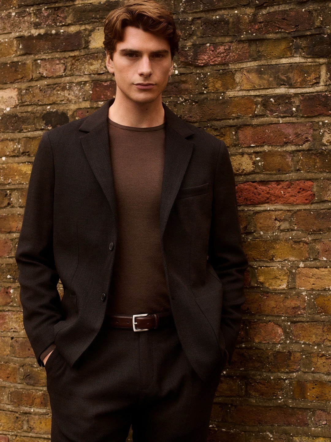 Dans le lookbook Sunspel, un homme porte un costume marron et une maille fine devant un mur en brique