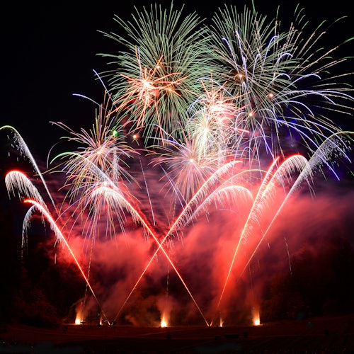 在黑夜的天空中,五颜六色的烟花以各种形状绽放,红色和白色的焰尾在下方的烟雾中闪耀。
