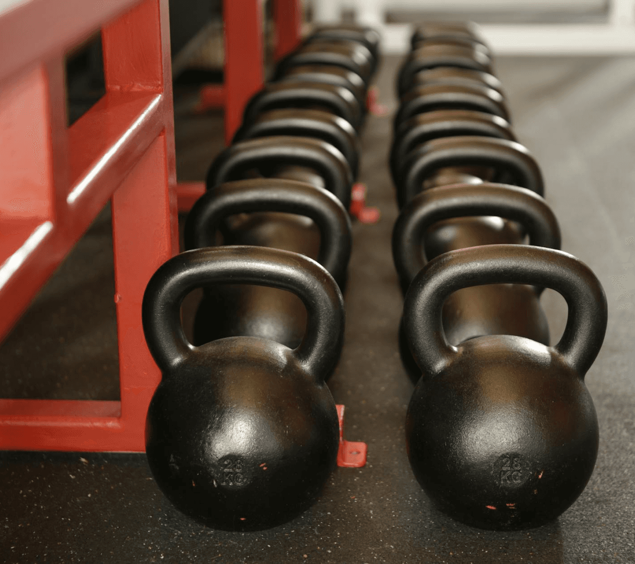 Two rows of black kettlebells on a dark gym floor next to a red metal rack. Front weights are marked 28 KG.