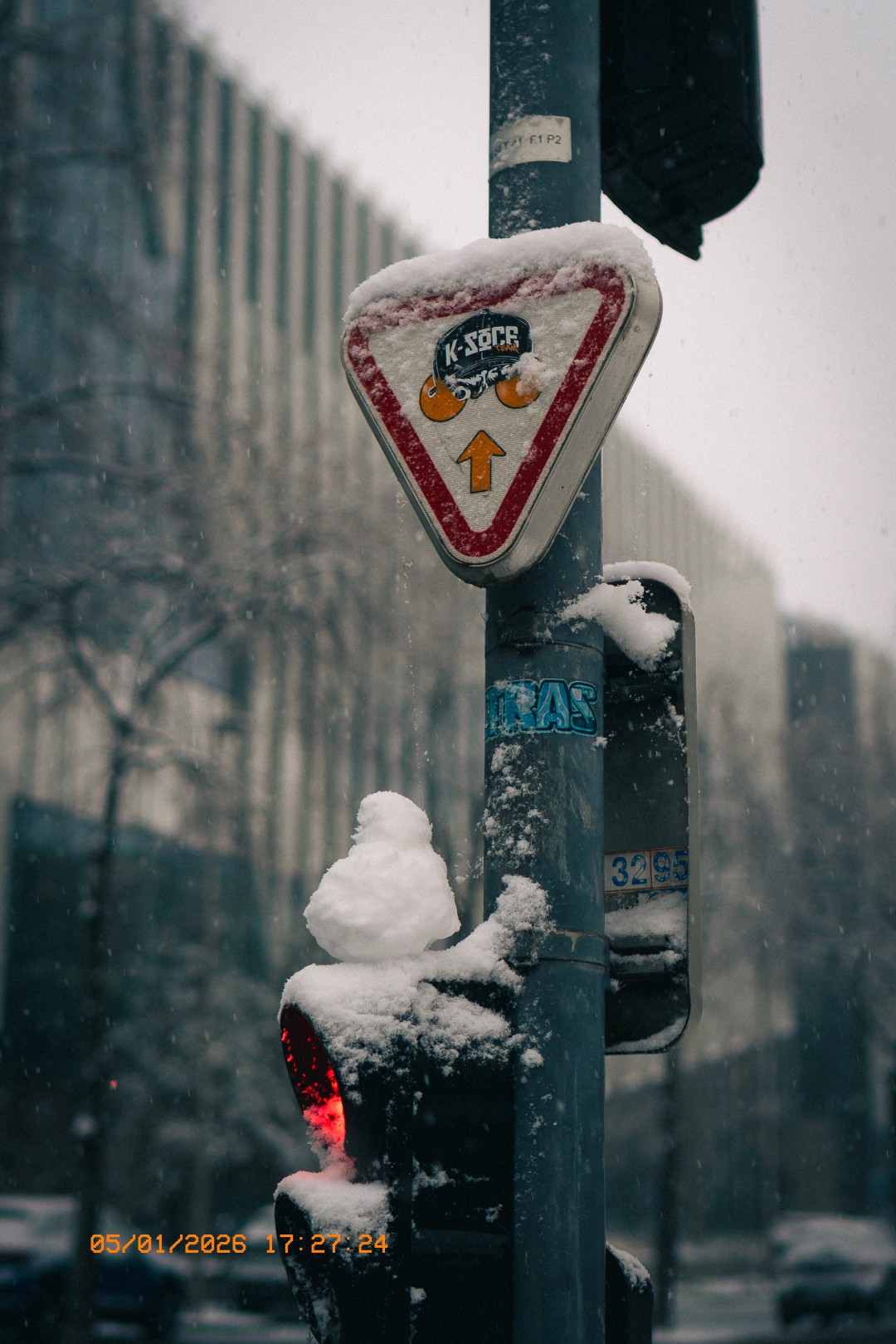 Signe de circulation enneigé avec un logo 'K-SOCC' et une flèche orange, sur un poteau de signalisation recouvert de neige, avec un bâtiment moderne en arrière-plan et une chute de neige dans une rue d'Issy.