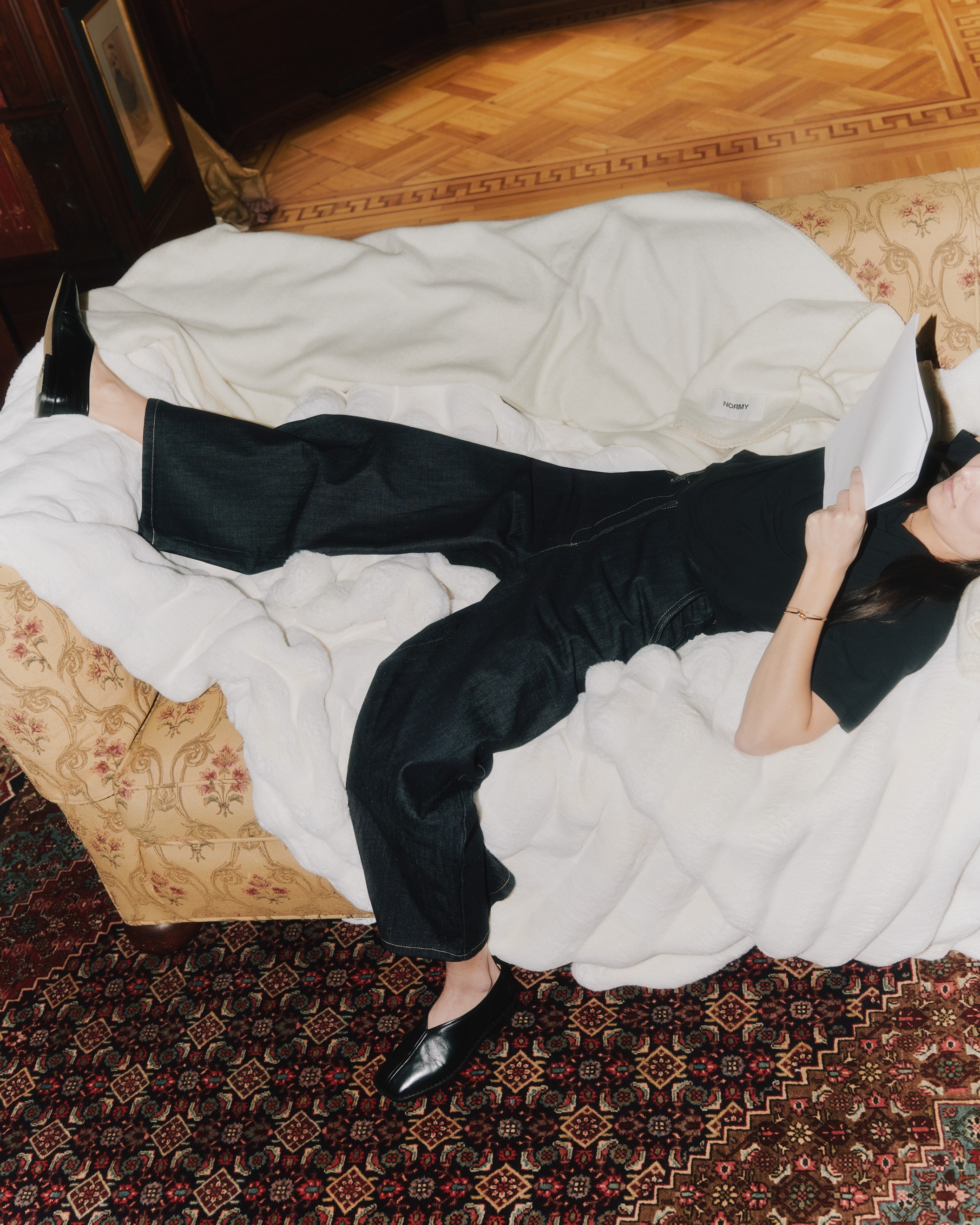 A cozy scene showing a person dressed in white lounging on a sofa, surrounded by plush blankets in cream and deep teal. Both blankets feature the "NORMY" label, highlighting the brand, and add a touch of comfort and style to the setting, which also features colorful tile flooring.