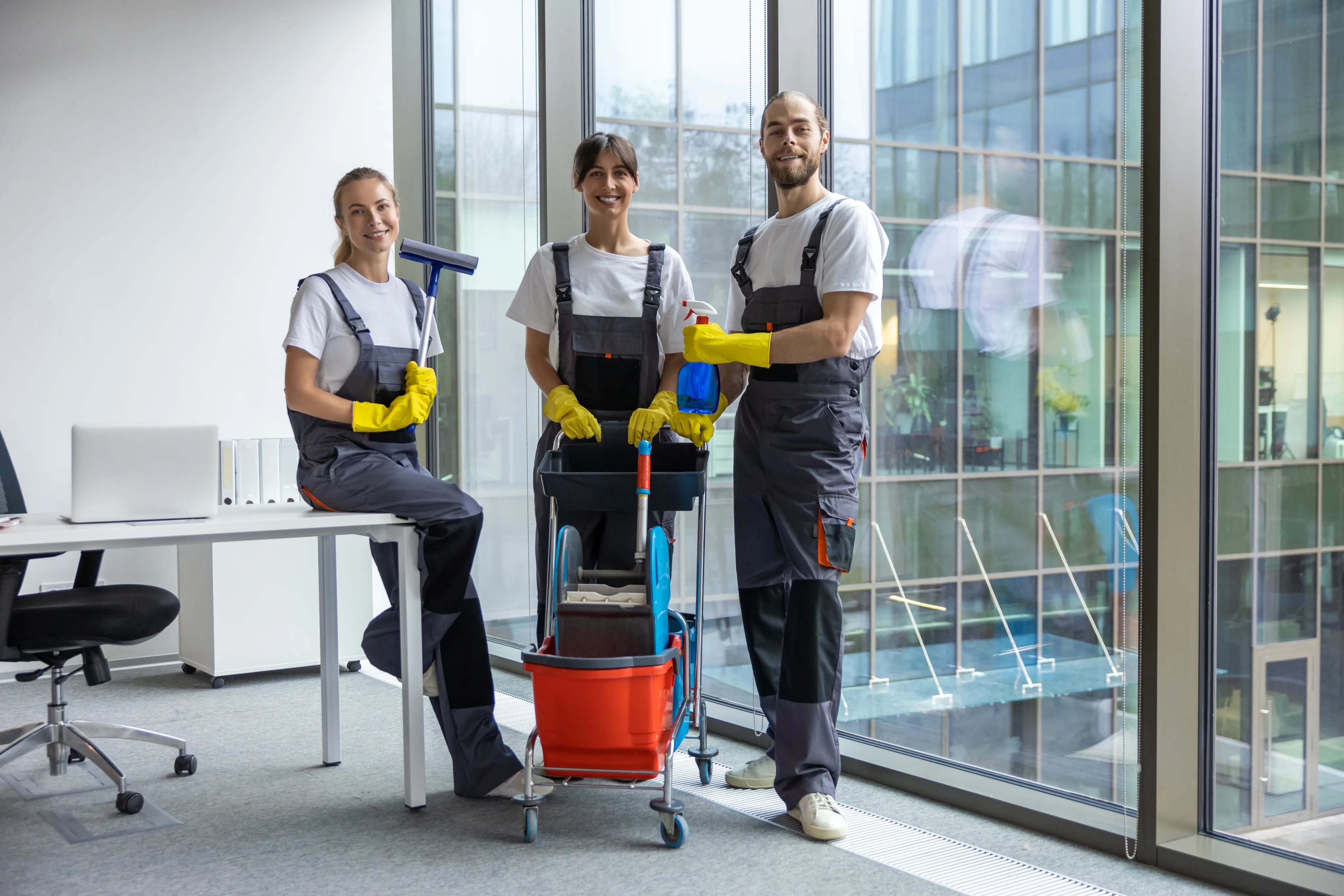 A diverse team of three professional cleaners in grey and yellow uniforms posing in a modern office setting with cleaning supplies.
