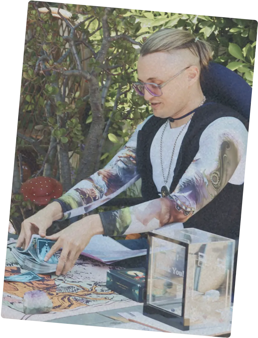 Person with with a pattern long-sleeve shirt, black vest, and glasses setting up a spiritual reading table with cards and crystals.
