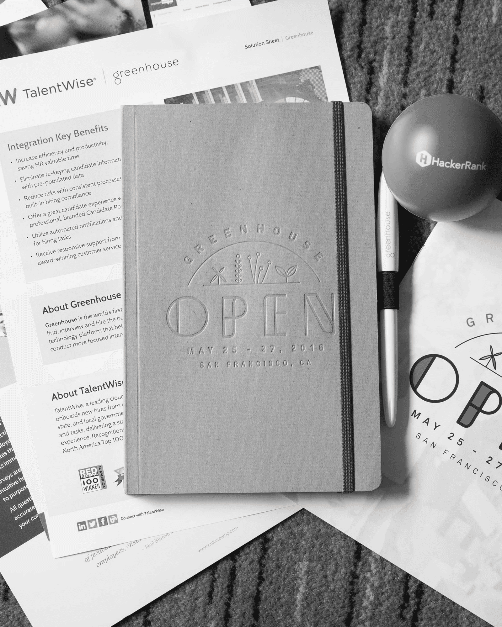 A flat lay of Greenhouse OPEN swag, a notebook, branded pen, and partner materials, documents the collateral produced for the rebrand.