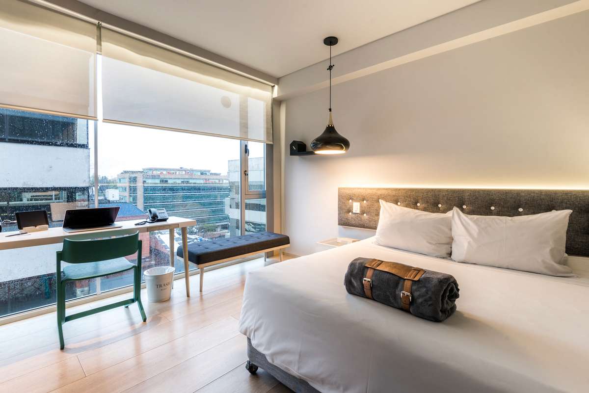 A modern hotel room with a large window overlooking a city street, featuring a bed with white linens and a desk chair.