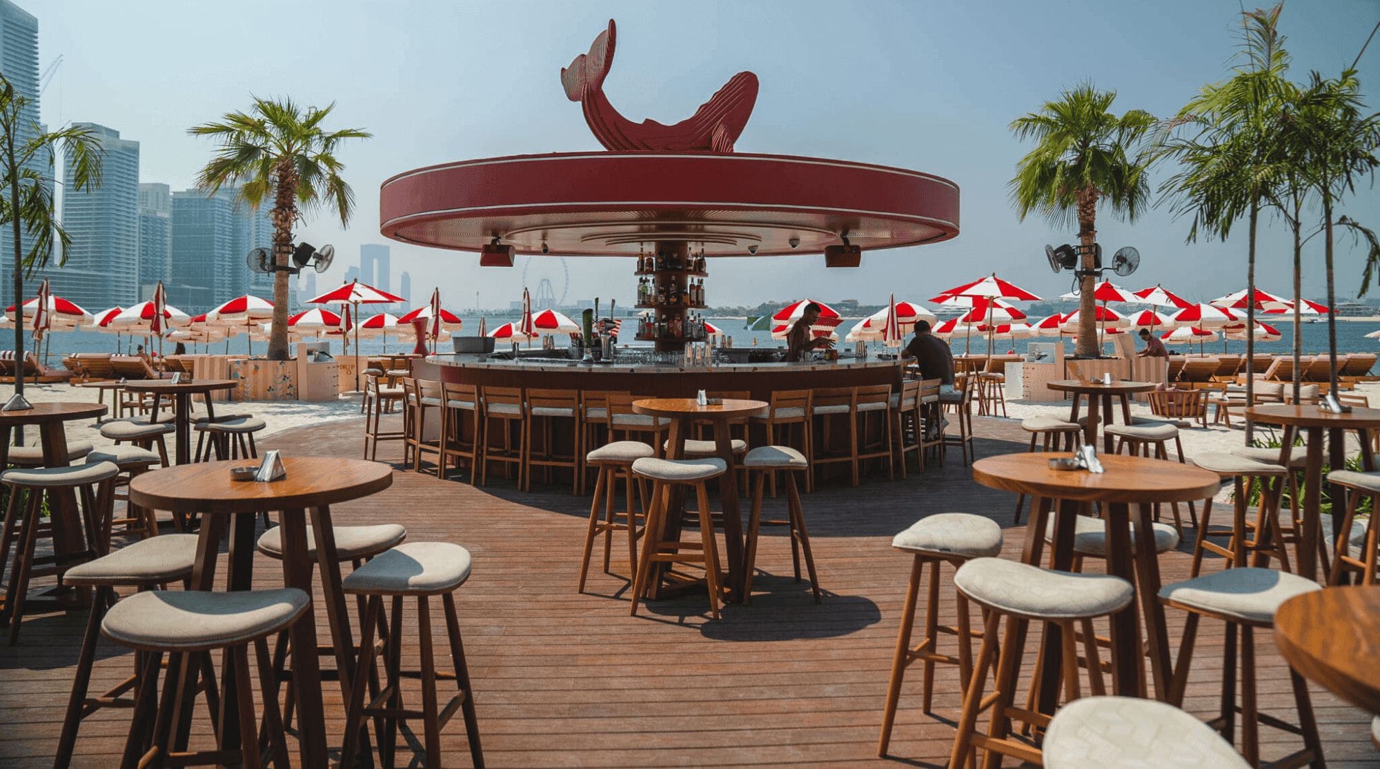 An outdoor beach restaurant in Dubai with a circular bar counter, wooden seating, and red umbrellas overlooking the sea.