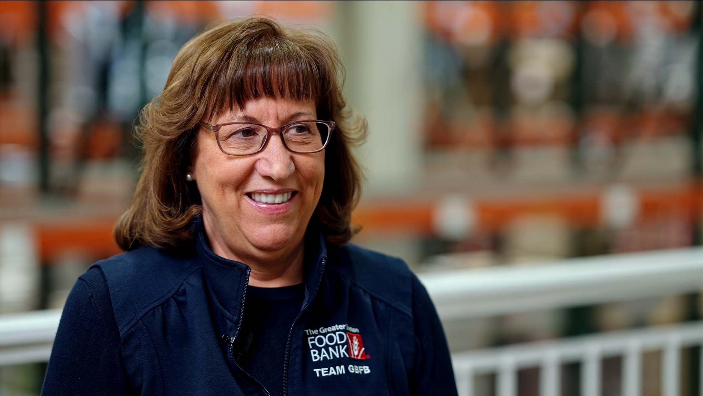 A woman wearing glasses smiles in a warehouse setting; the logo on her vest reads “The Greater FOOD BANK” and “TEAM GFB.”