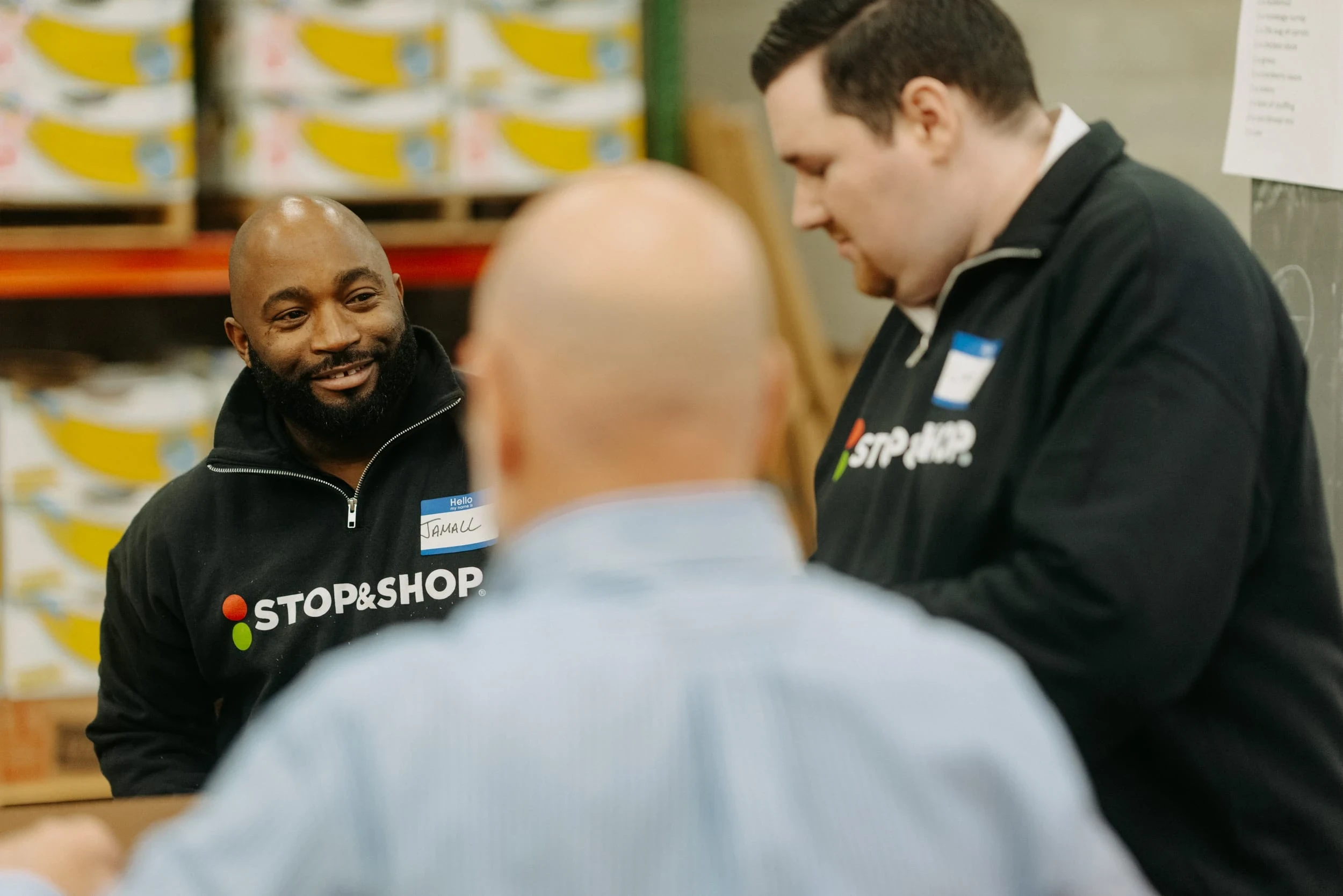 A close-up, candid photograph taken indoors, likely in a warehouse or stock room, showing three men gathered around a table. A Black man with a beard, identified by his name tag as Jamall, is on the left, smiling and wearing a black Stop & Shop branded hoodie. A white man to his right is also wearing a dark shirt with the Stop & Shop logo and is looking down. In the immediate foreground, the back of a third person's head and shoulders, wearing a light-colored shirt, is out of focus. Pallets and stacked boxes are visible on shelves in the background.