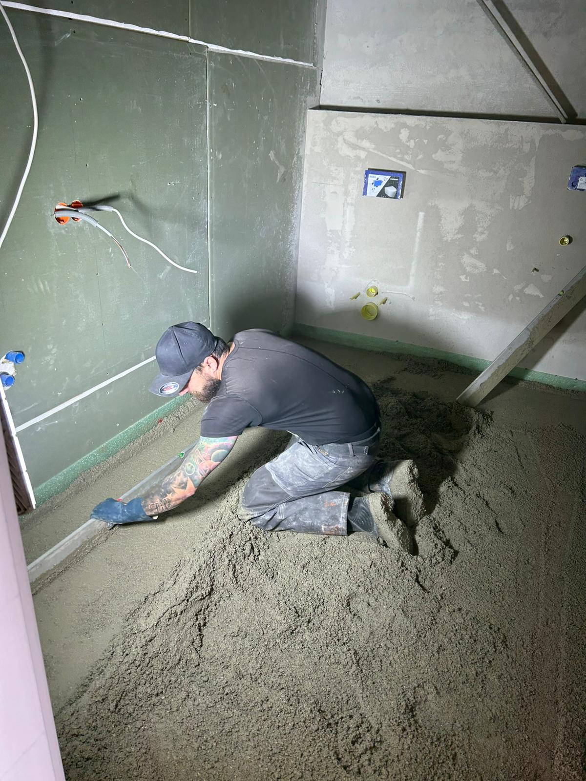 Ein Bauarbeiter kniet und glättet während einer Zimmerrenovierung mit einem langen Werkzeug den Betonboden.
