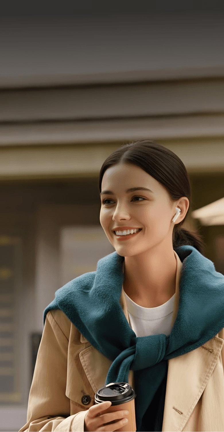 Smiling woman with earbuds, wearing a blue scarf and trench coat, holding coffee, walking on a city street.