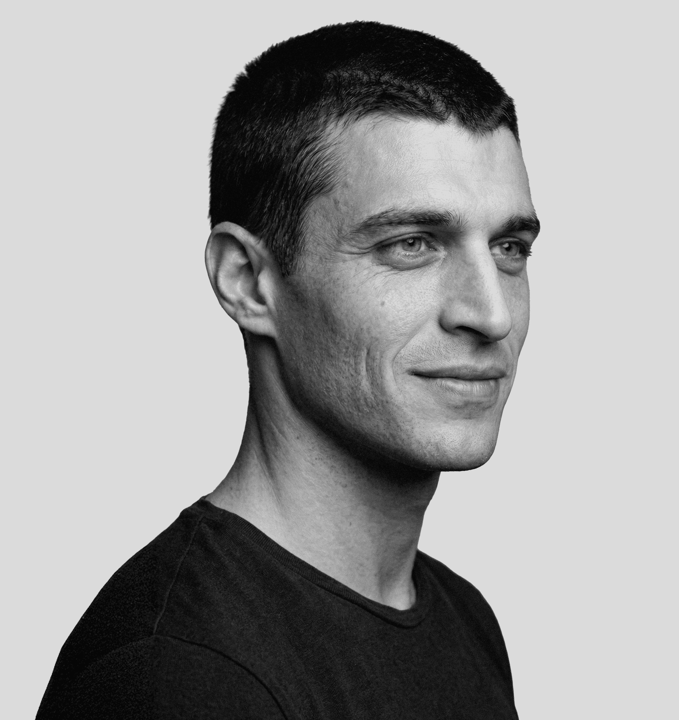 Portrait of a man with short dark hair and a trimmed beard, dressed in a dark shirt, on a simple background.