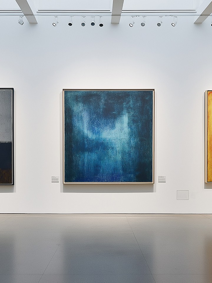 Minimalist gallery interior shot showing three large framed abstract colour-field paintings (charcoal, deep blue, and orange-yellow) hung on a white wall under overhead spotlights.