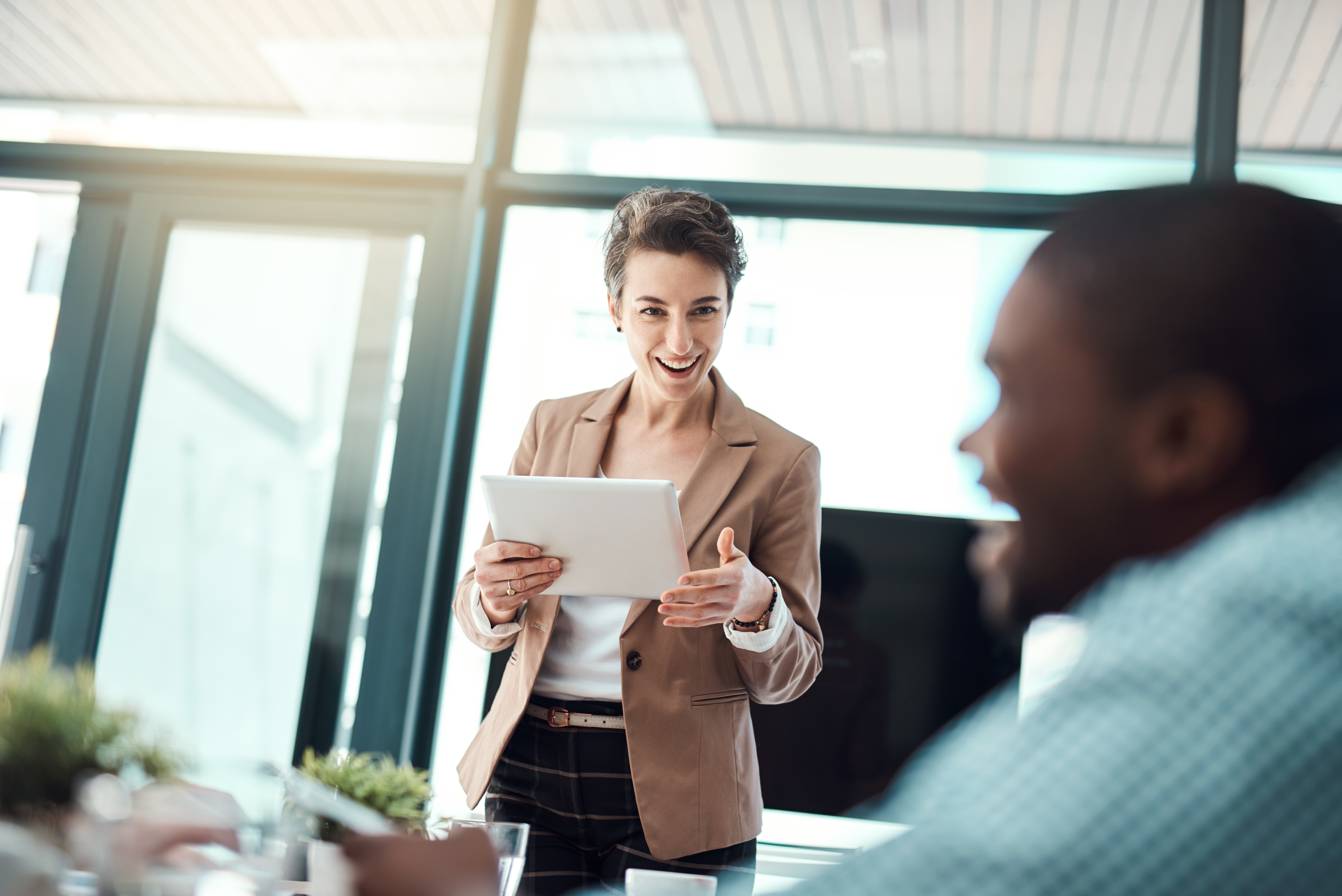 Lächelnde Geschäftsfrau in heller Bluse und Blazer hält ein Tablet und spricht mit Kolleg:innen in einem modernen, hellen Büro.