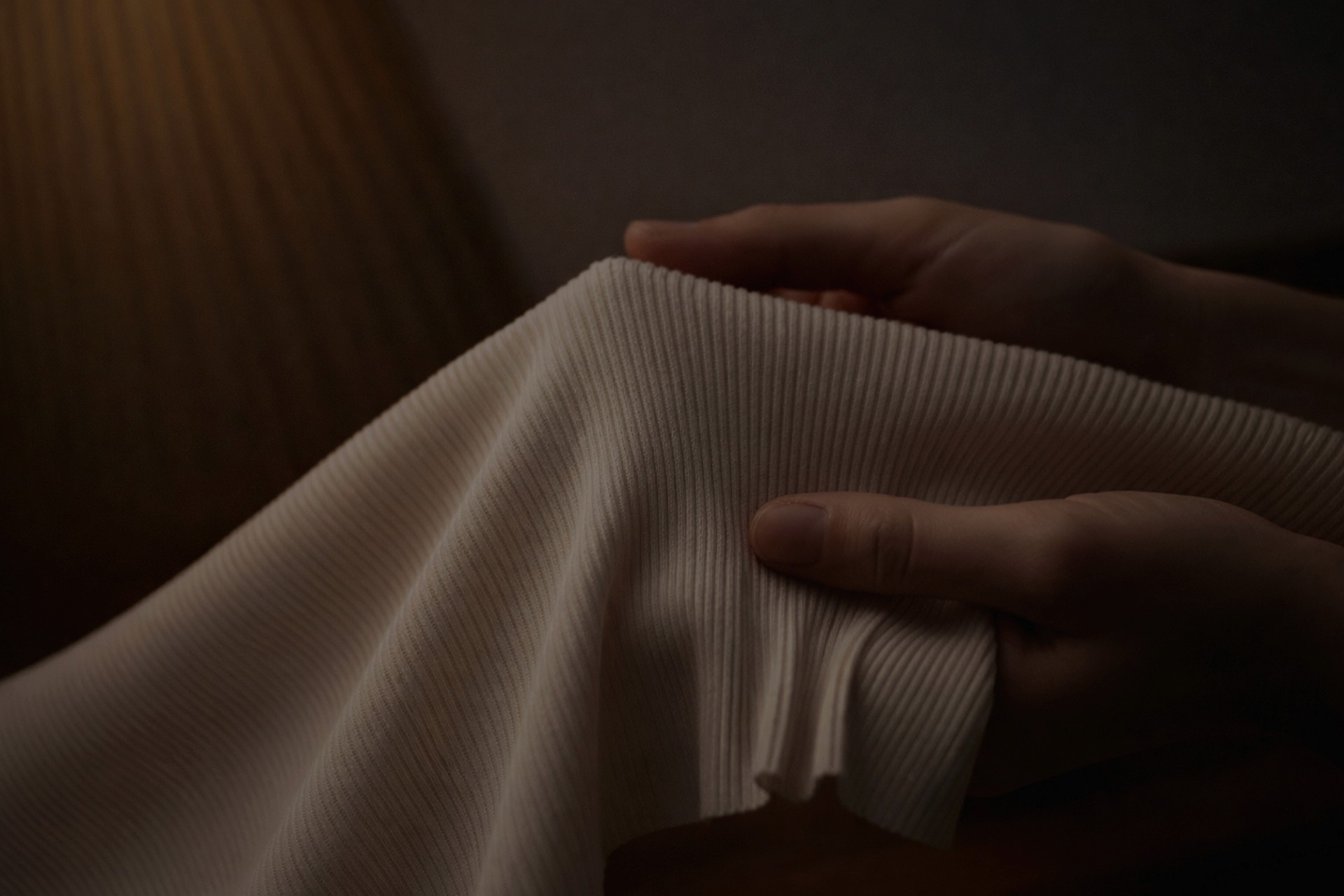 A close-up of hands holding and stretching a piece of creamy, fine-ribbed fabric. The texture of the material is highlighted by a soft, warm light in a dimly lit setting.
