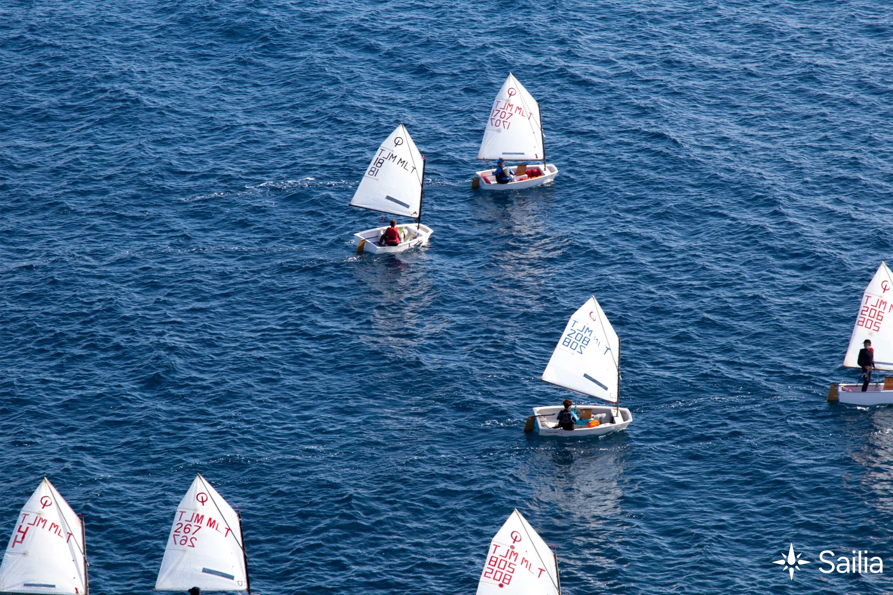 A group of small sailboats with white sails on a deep blue sea arranged in a scattered pattern. Each boat has a single person, creating a sense of motion and calm.