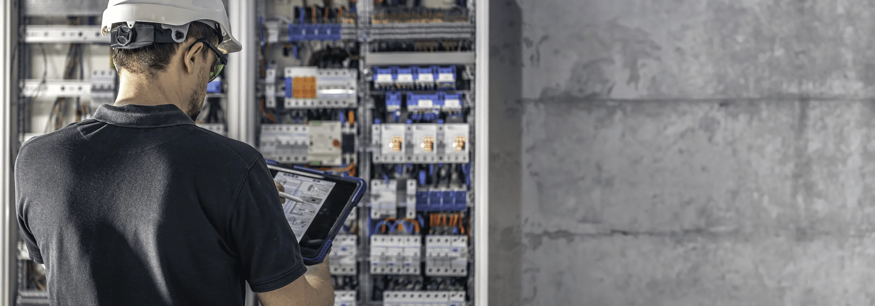 Ein Techniker mit Schutzbrille bedient das Tablet vor einem geöffneten Schaltschrank voller Kabel.