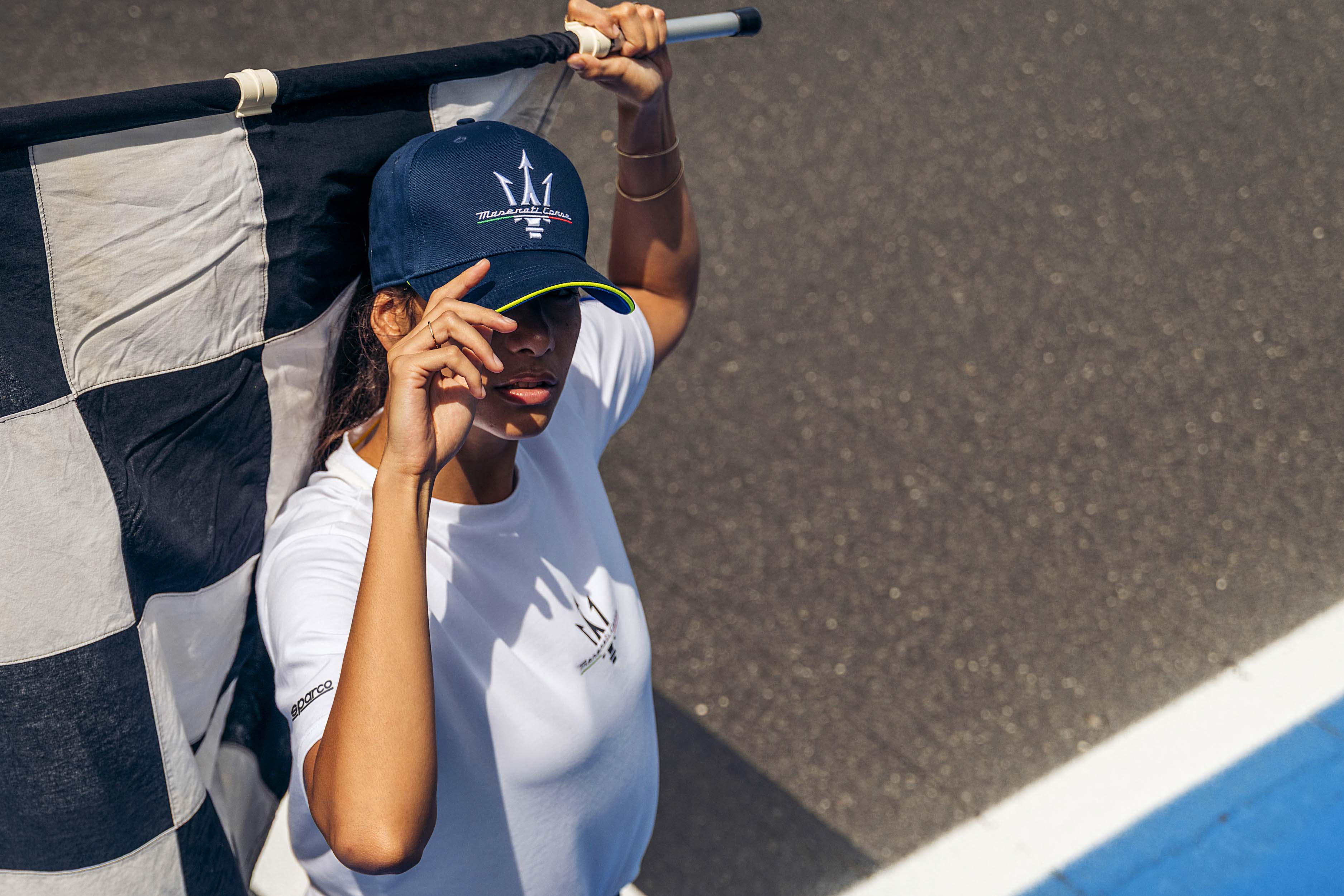 Femme portant une casquette Maserati bleu marine et un t-shirt blanc, posant sur un circuit de course avec un drapeau à la main.