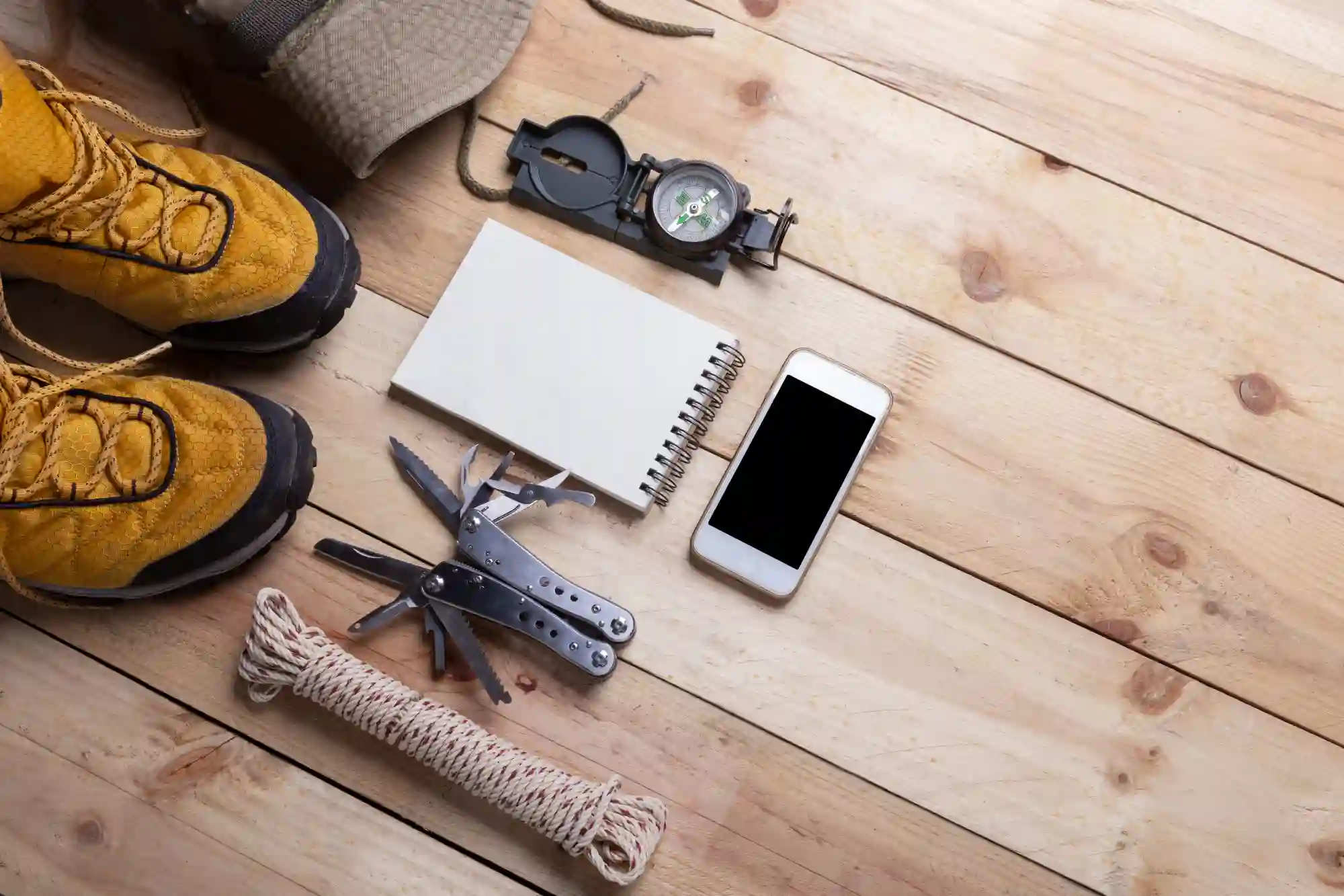Hiking gear on wood: boots, compass, rope, knife, phone.