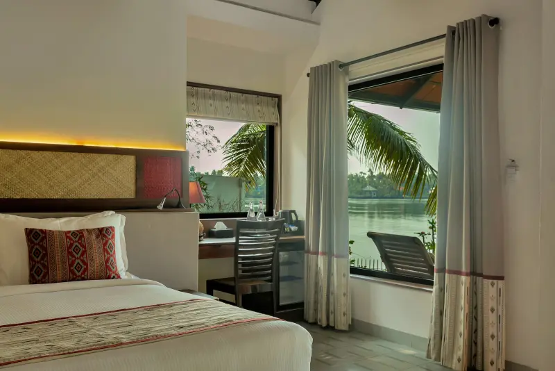 Lake View Villa bedroom at Vedic Village Kodungallur with a king bed, writing desk and floor-to-ceiling window overlooking the lake and coconut palms