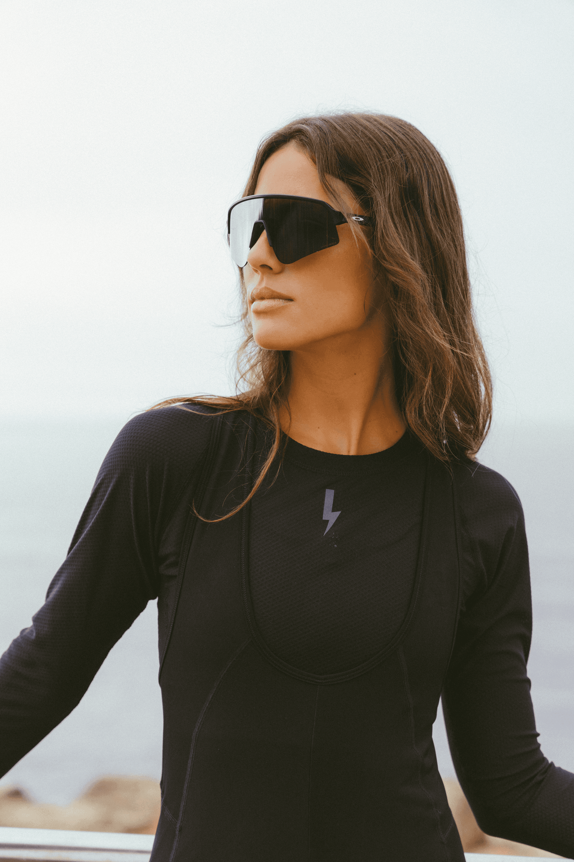 A-portrait-shot-of-a-young-woman-with-long-dark-wavy-hair-wearing-a-black-long-sleeve-base-layer-shirt-with-a-subtle-mesh-She-also-wearing-large-sleek-black-wraparound-sports-sunglasses-and-is-looking-to-the-right-The-background-is-a-slightly-blured-foggy-view-over-the-ocean-The-lighting-bolt-sign-is-visible-on-the-chest-of-the-base-layer
