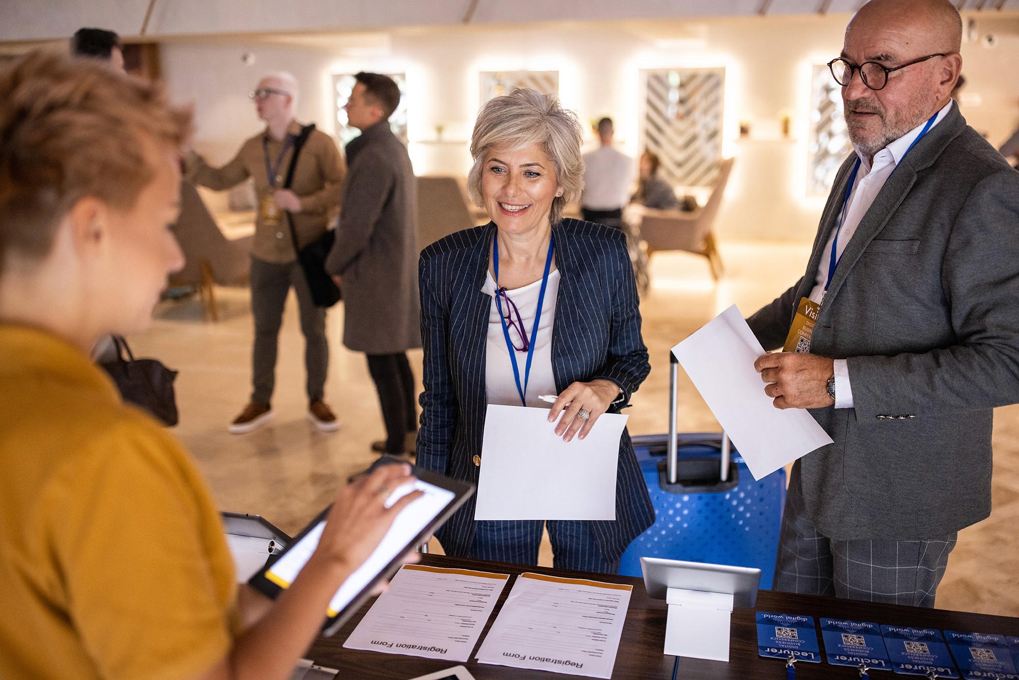 People at a conference registration desk, holding papers and talking to staff with a tablet.