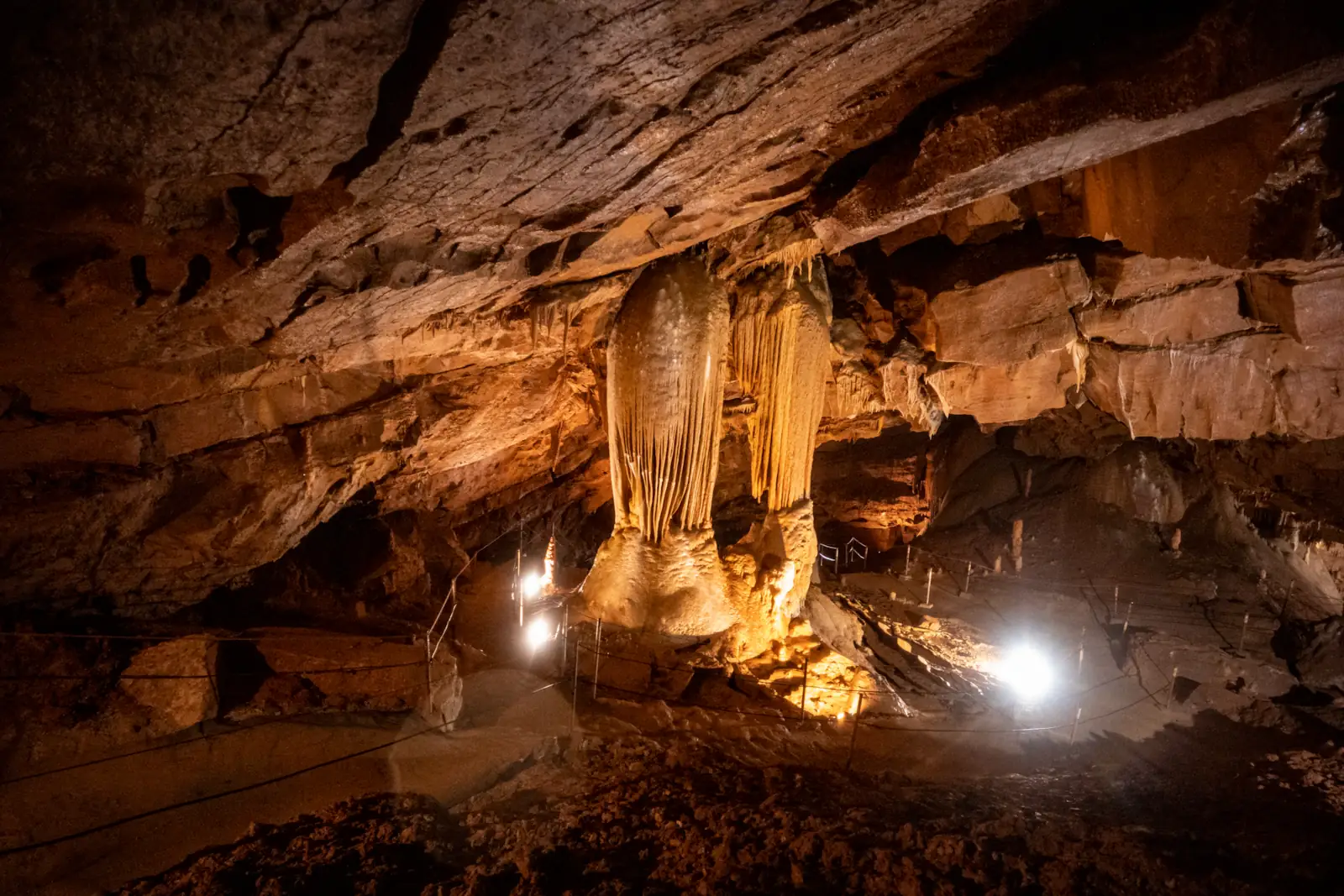 Interior of Mayor's Cave in Slovenia, featuring massive limestone stalactites and stalagmites illuminated by warm spotlights along a fenced tourist path deep underground.