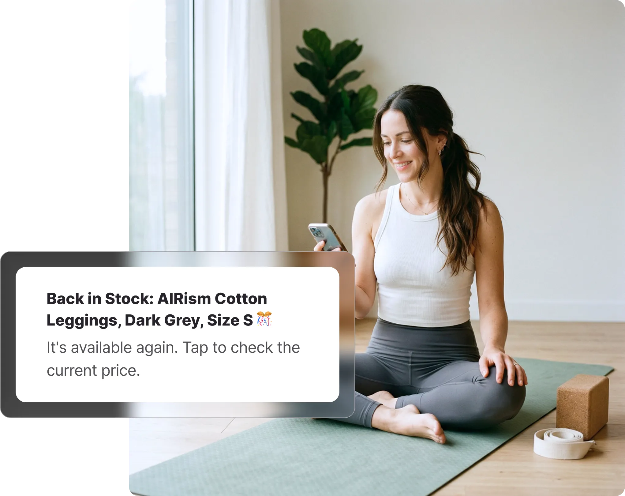 Woman sitting cross-legged on a yoga mat checking her phone, receiving a Uniqlo back-in-stock notification on Whisprice, with a cork yoga block and strap beside her.