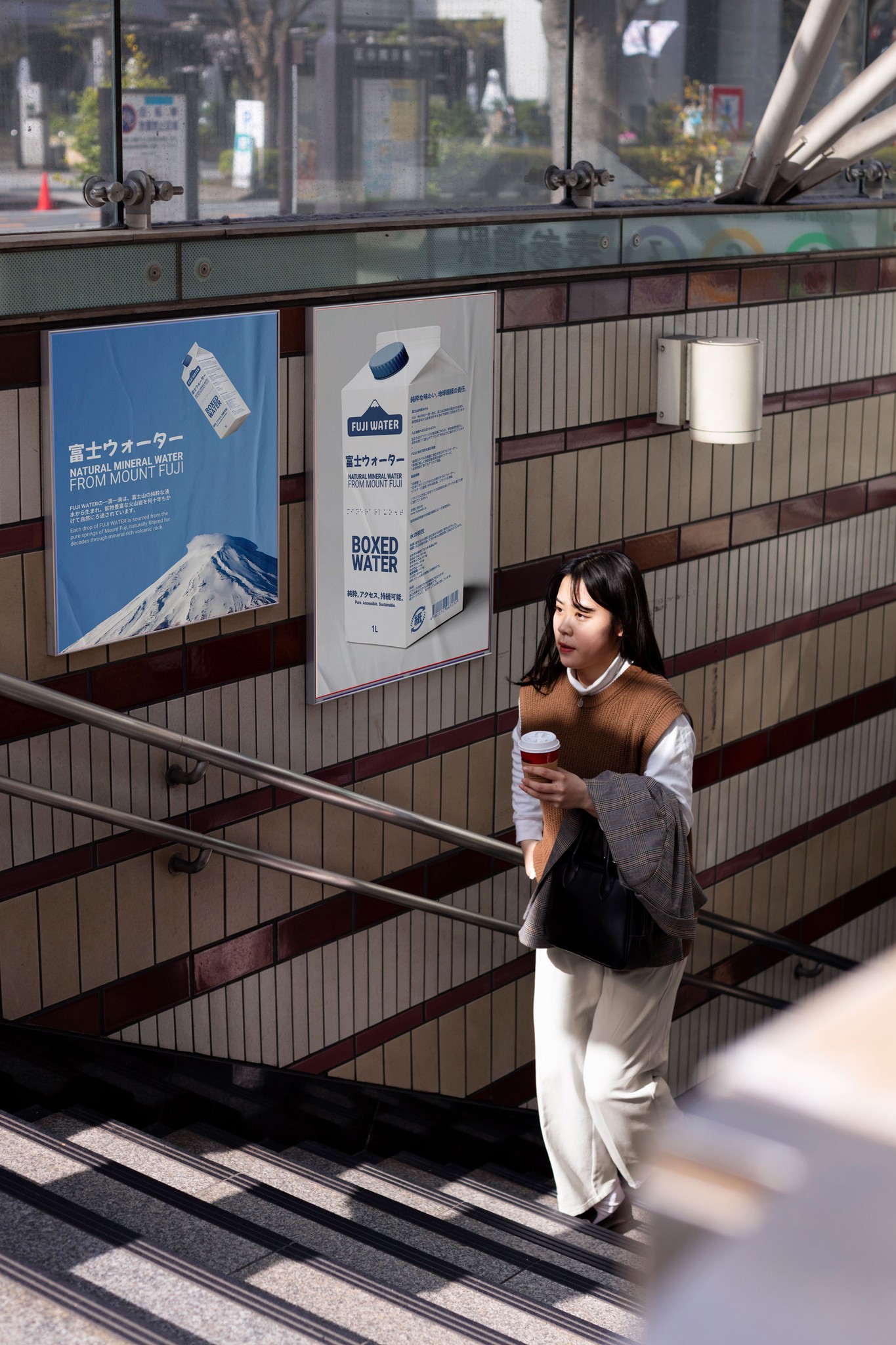 A person stands near a wall with posters, next to stairs, in a modern urban setting. Bright daylight is visible.