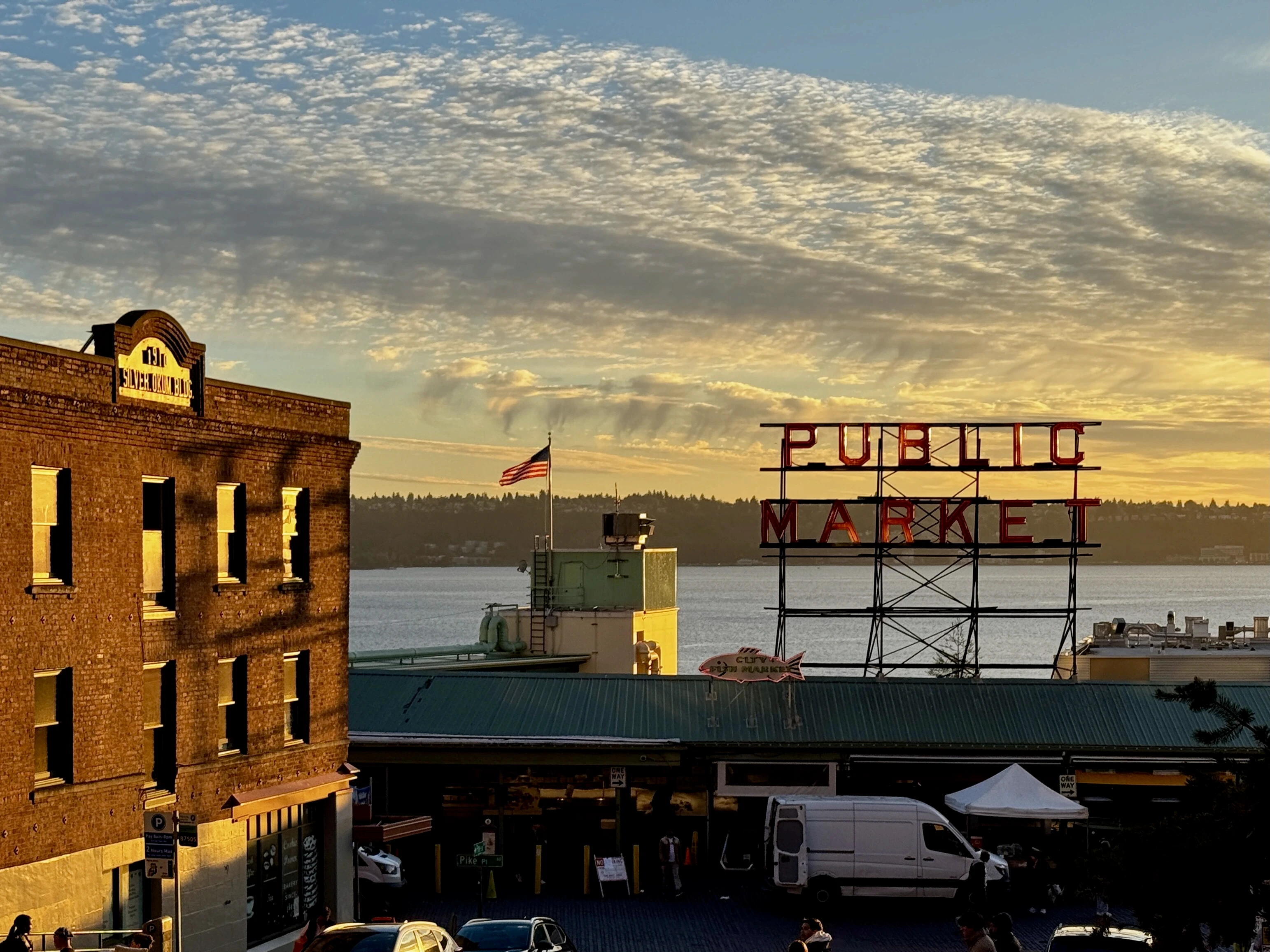 The Pike Place Market sign glows in warm evening light above Seattle’s waterfront, with Elliott Bay visible in the background.