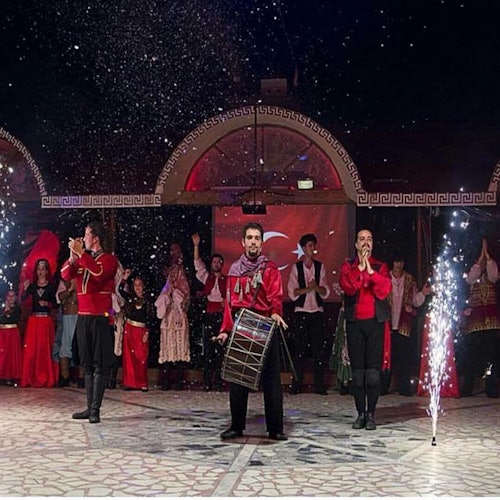 Artister i röda kläder står på en scen med konfetti som faller och gnistor som tänds, medan publiken applåderar i bakgrunden.