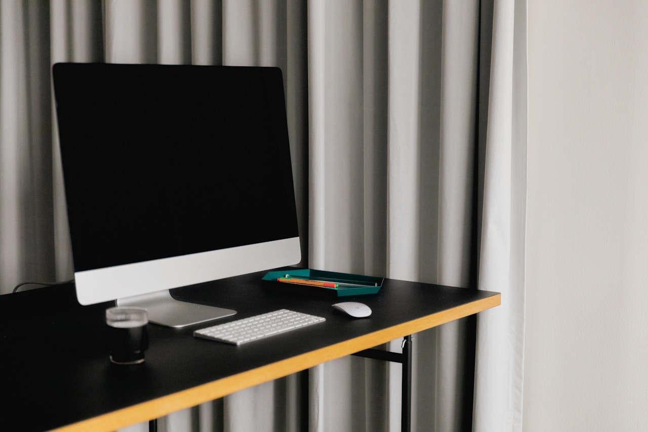 Modern desktop computer with blank screen on a black desk, accompanied by a keyboard, mouse, pencil tray, and curtain backdrop, symbolizing digital tools for laundromat management.