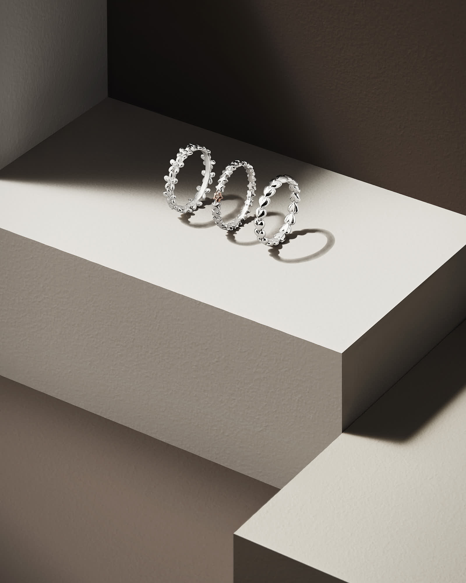 Three simple metallic rings displayed on a minimalistic light-colored pedestal against a brown background.