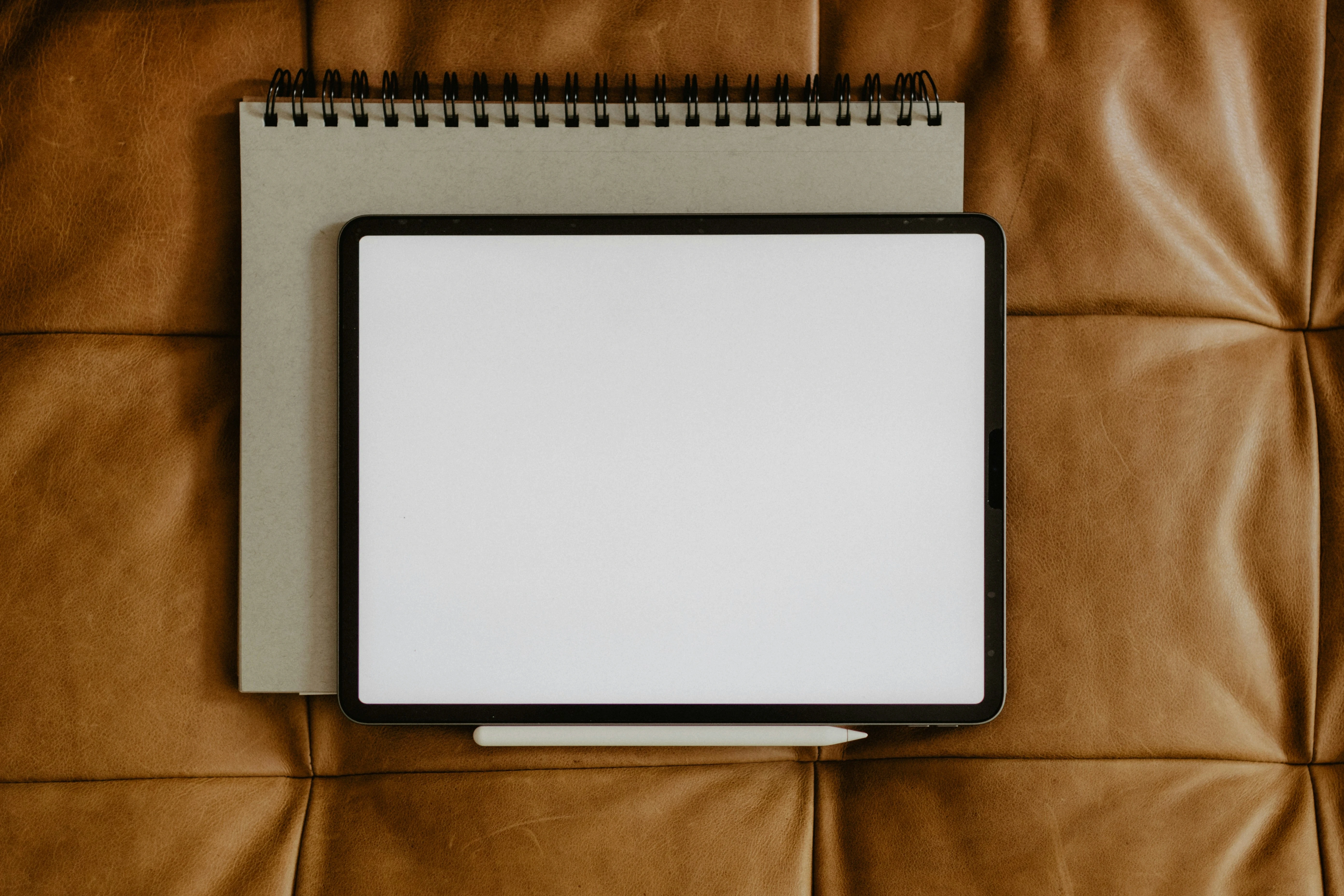 iPad with Apple Pencil placed over a spiral-bound notebook on a brown leather surface.