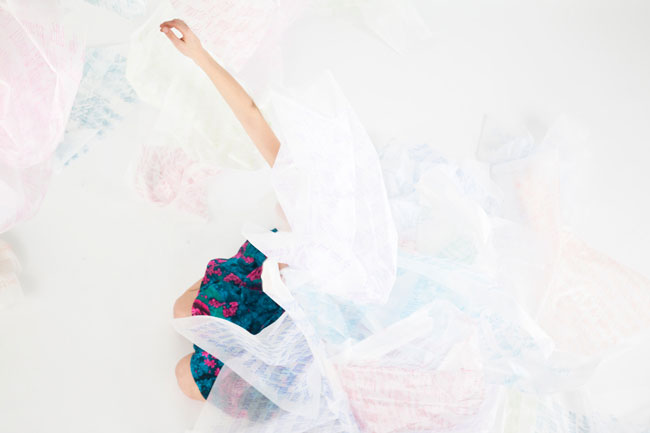 A person in a teal floral dress is positioned upside down on a white surface, with their right arm extended upward and left arm bent at the elbow. A translucent white fabric drapes over the person’s head and upper body. The fabric has faint pink and blue markings. The person’s left arm is partially obscured by the fabric. The background is entirely white.