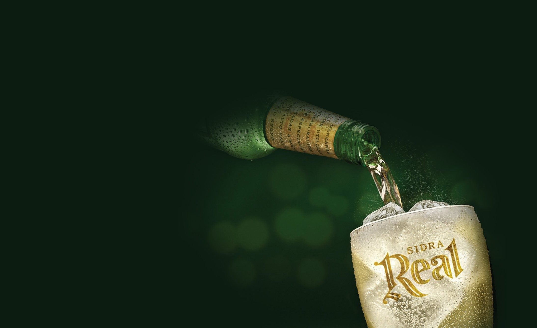 A green bottle pours golden cider into an icy glass labeled "Sidra Real" against a dark background, evoking a refreshing and crisp feeling.