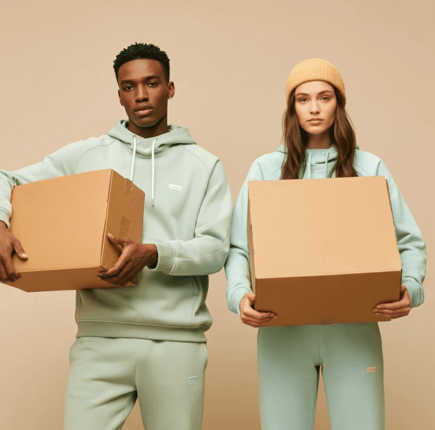 One male and one female fashion model holding cardboard boxes of clothes in a magazine cover style, both wearing athletic plain clothing with neutral tones 