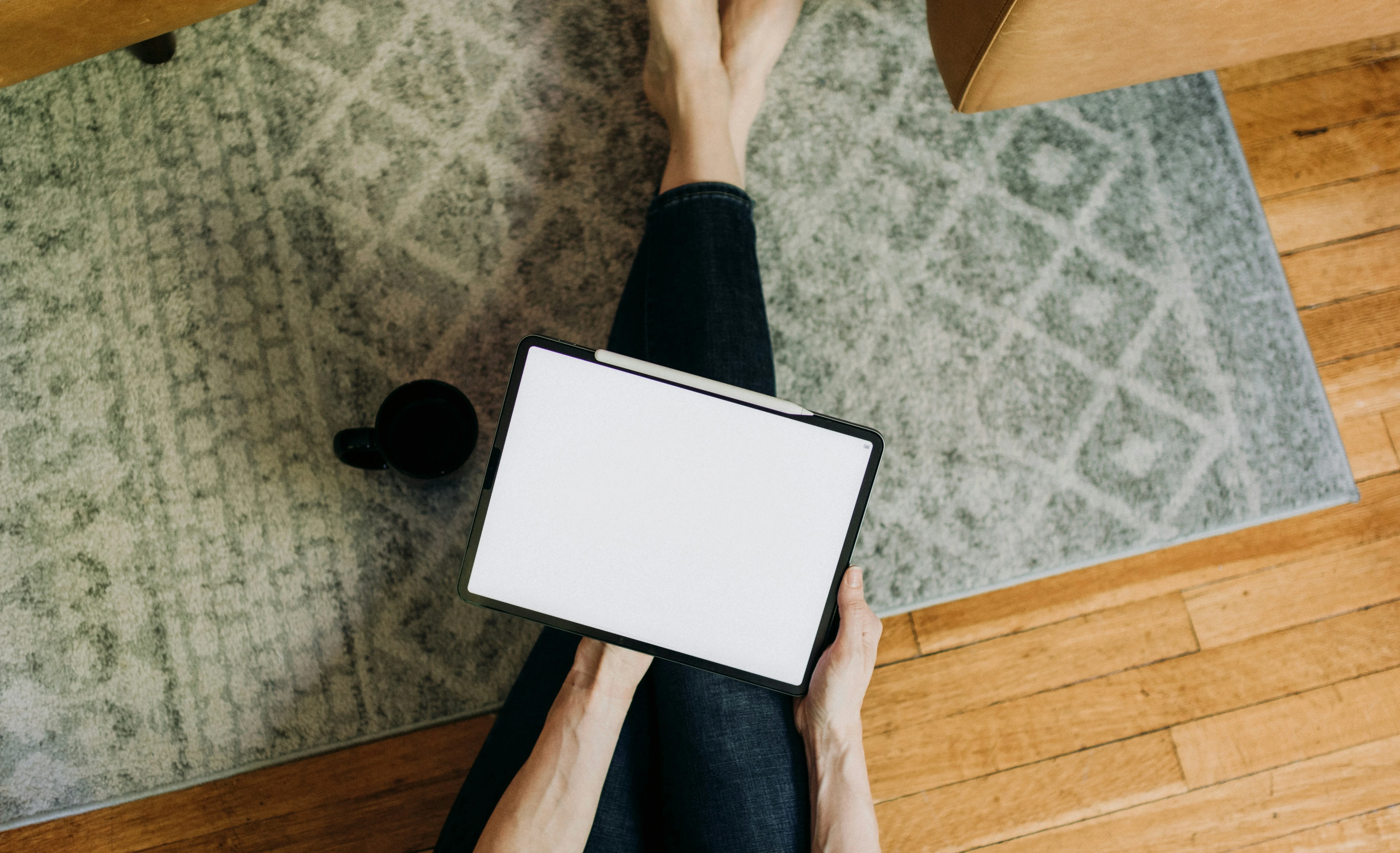 Person sitting cross-legged on a rug, holding an iPad with Apple Pencil resting on top, coffee mug nearby.