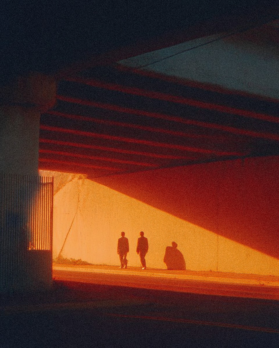 Two people walk beneath an overpass where bright orange sunlight hits a concrete wall, casting long, distinct shadows in the dim underpass.