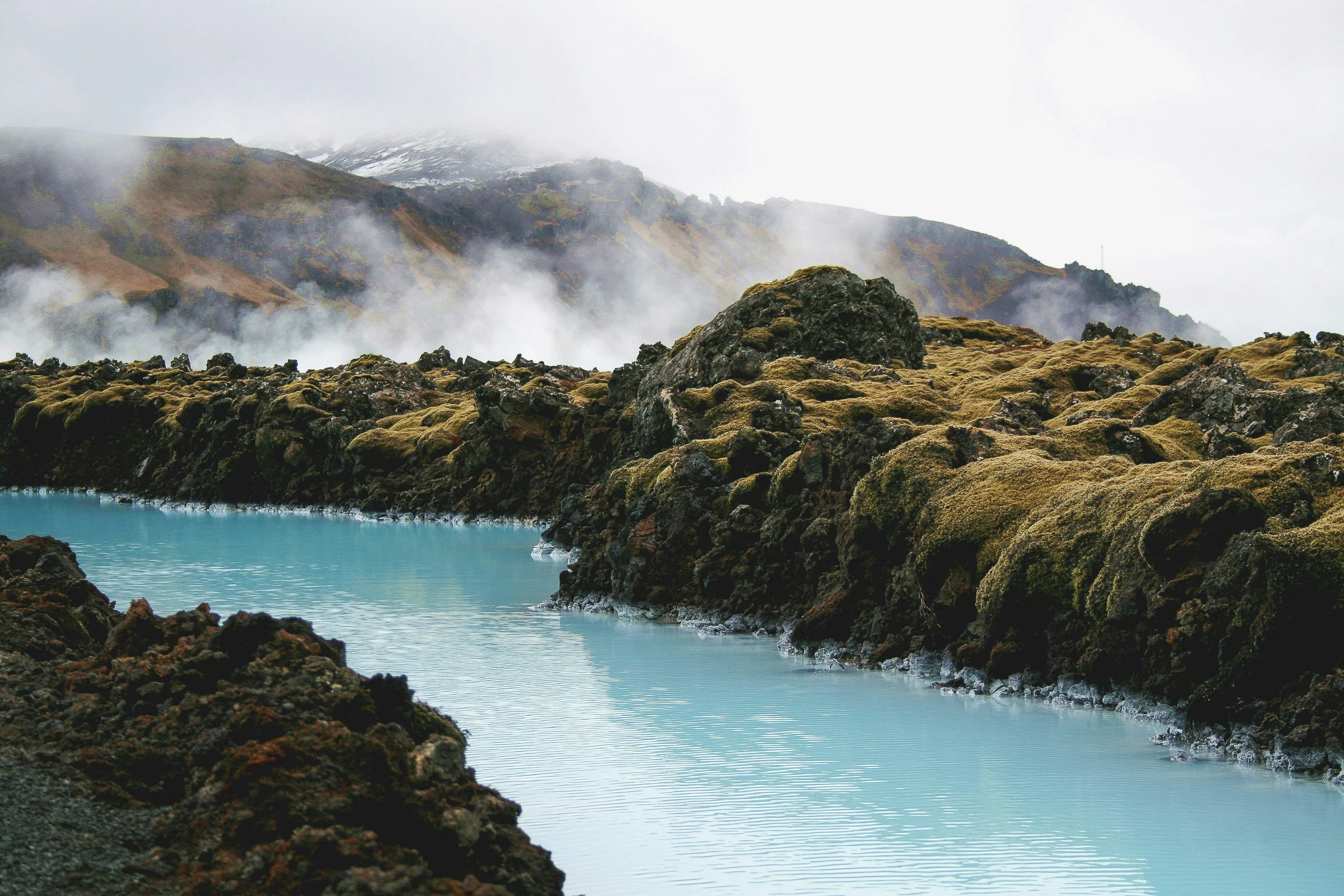 A milky blue geothermal pool surrounded by dark, moss-covered lava rocks with steam rising from the surface.