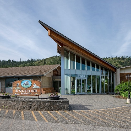 建築入口有一個三角形屋頂、大型玻璃窗，還有一個寫著「BC 野生動物公園甘露市」的標誌。背景是山和天空。