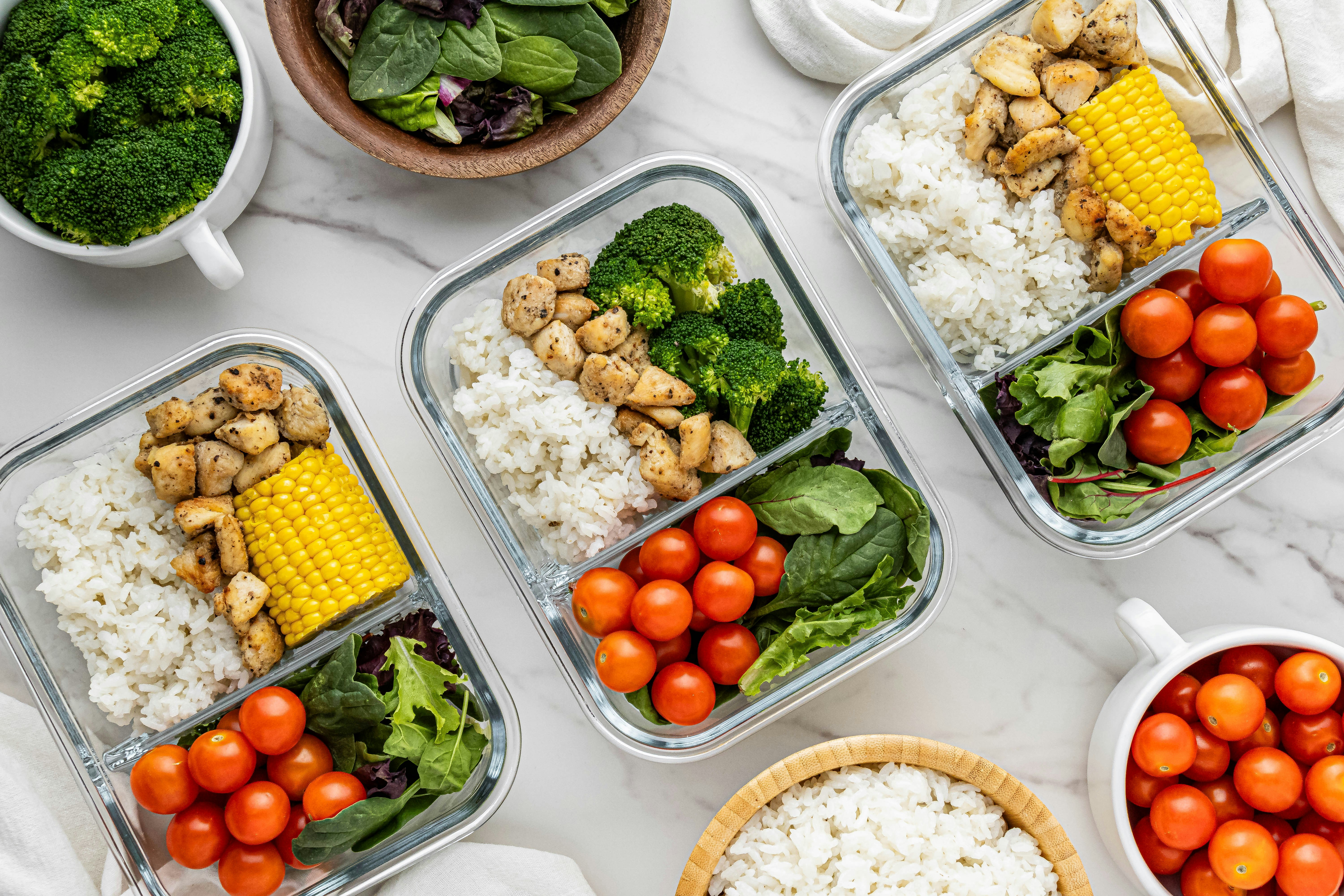 Meal prep containers with rice, chicken, vegetables, and cherry tomatoes on a white countertop.