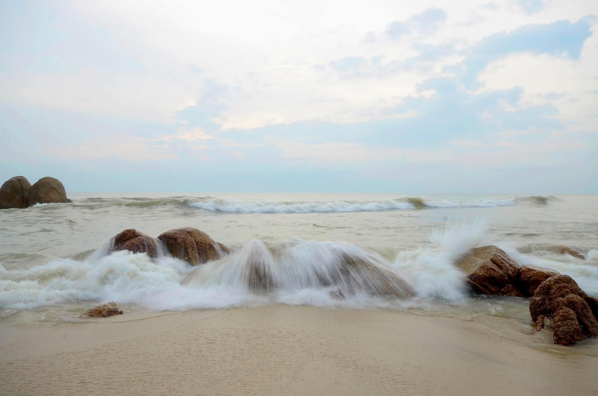 Gentle waves crash over smooth rocks on a sandy beach under a cloudy sky with soft, muted light, conveying tranquility and calmness.