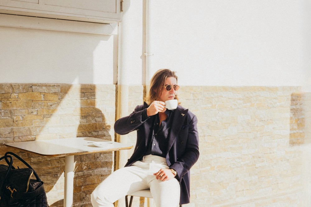 Stylish man enjoying coffee at an outdoor café, wearing a navy blazer, white trousers, and sunglasses under warm sunlight, reflecting refined luxury and effortless confidence.