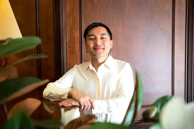 Uma pessoa vestindo uma camisa branca está sentada com confiança em uma sala com iluminação acolhedora, com painéis de madeira e plantas verdes frondosas em primeiro plano, transmitindo uma sensação de profissionalismo e tranquilidade.