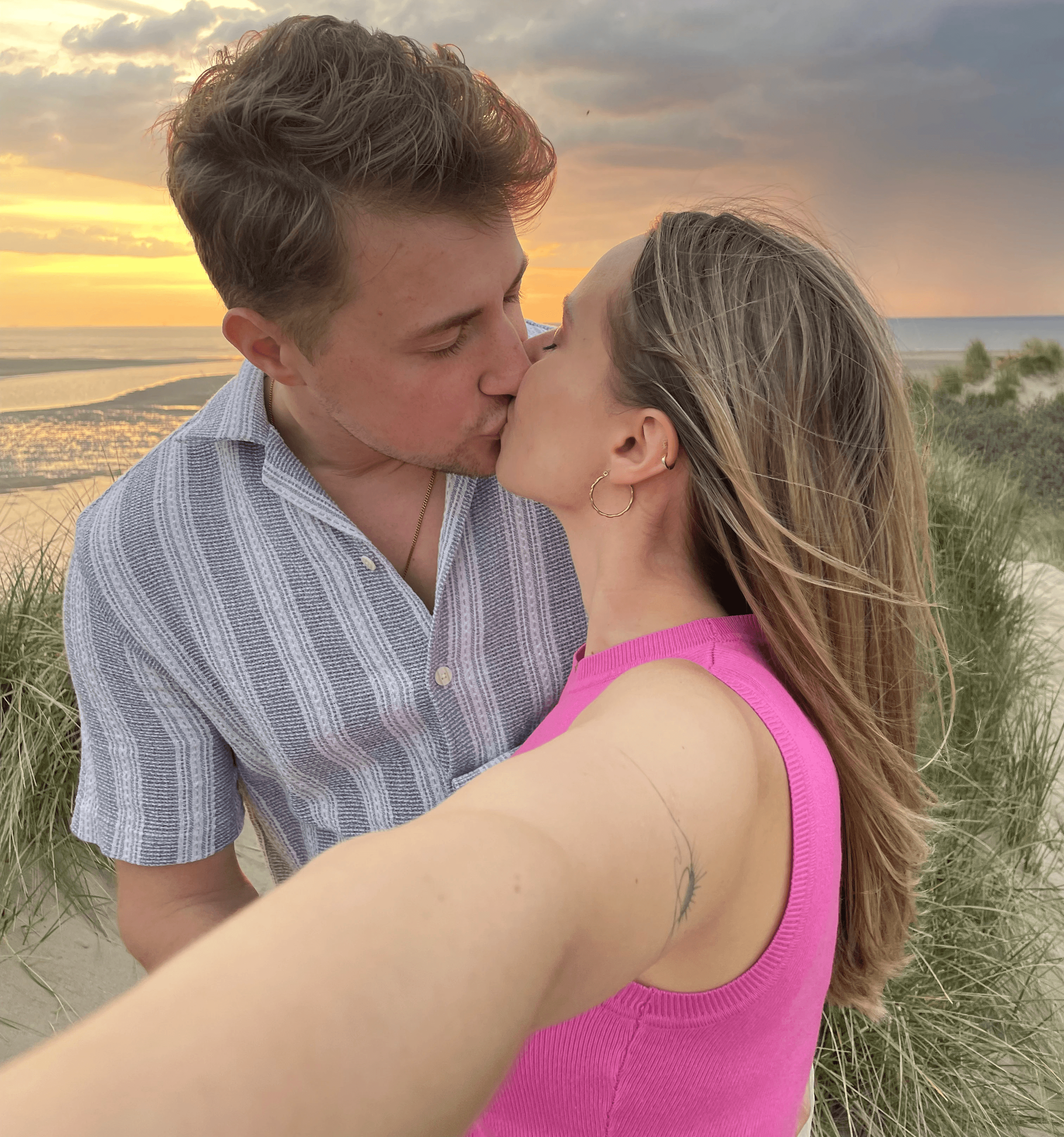 A couple holds hands, standing on a rocky beach under a large cave arch, gazing at the tranquil ocean during sunset. The sky is softly lit in pastel hues.